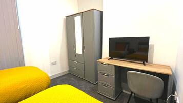 A hotel room with two yellow beds, a grey wardrobe, a desk with a television, and a grey chair.