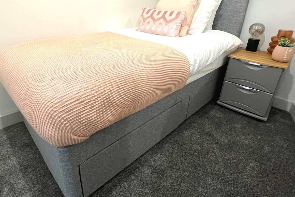 A neatly made bed with a grey headboard, a grey nightstand with drawers, and a small potted plant on the nightstand.