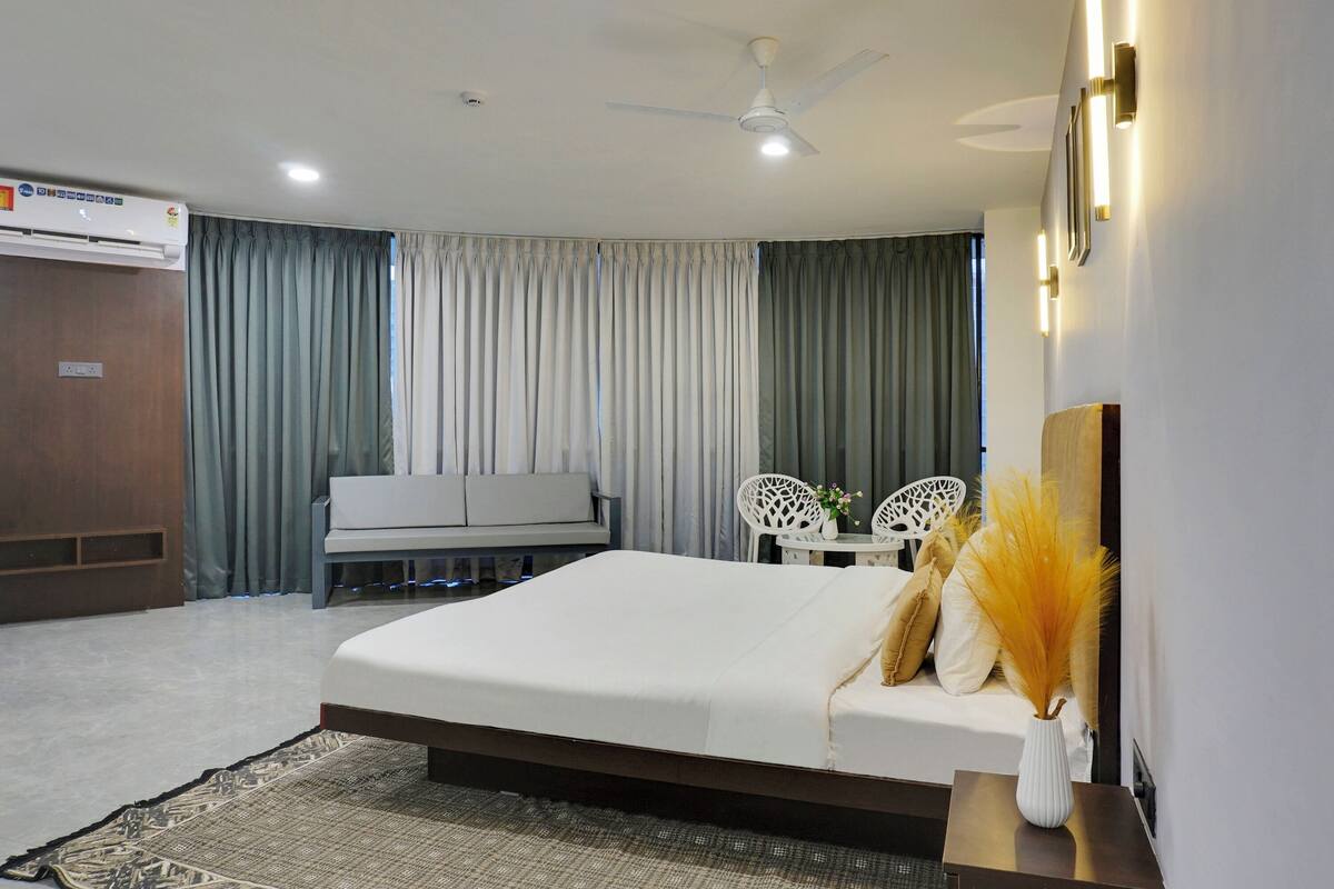 A hotel room with a bed, a sofa, a small table with chairs, an air conditioning unit, and a ceiling fan.