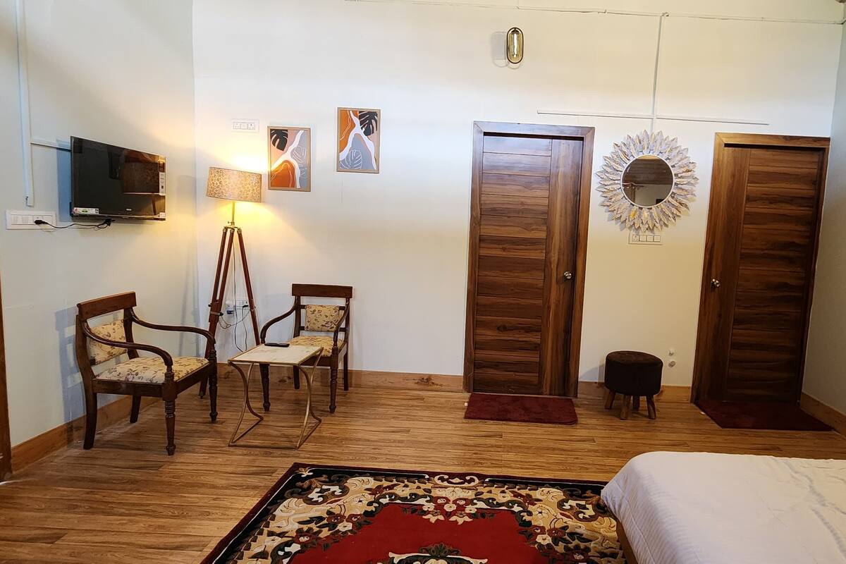 A bedroom with a bed, two wooden wardrobes, a television mounted on the wall, two chairs, a small stool, and a patterned rug.