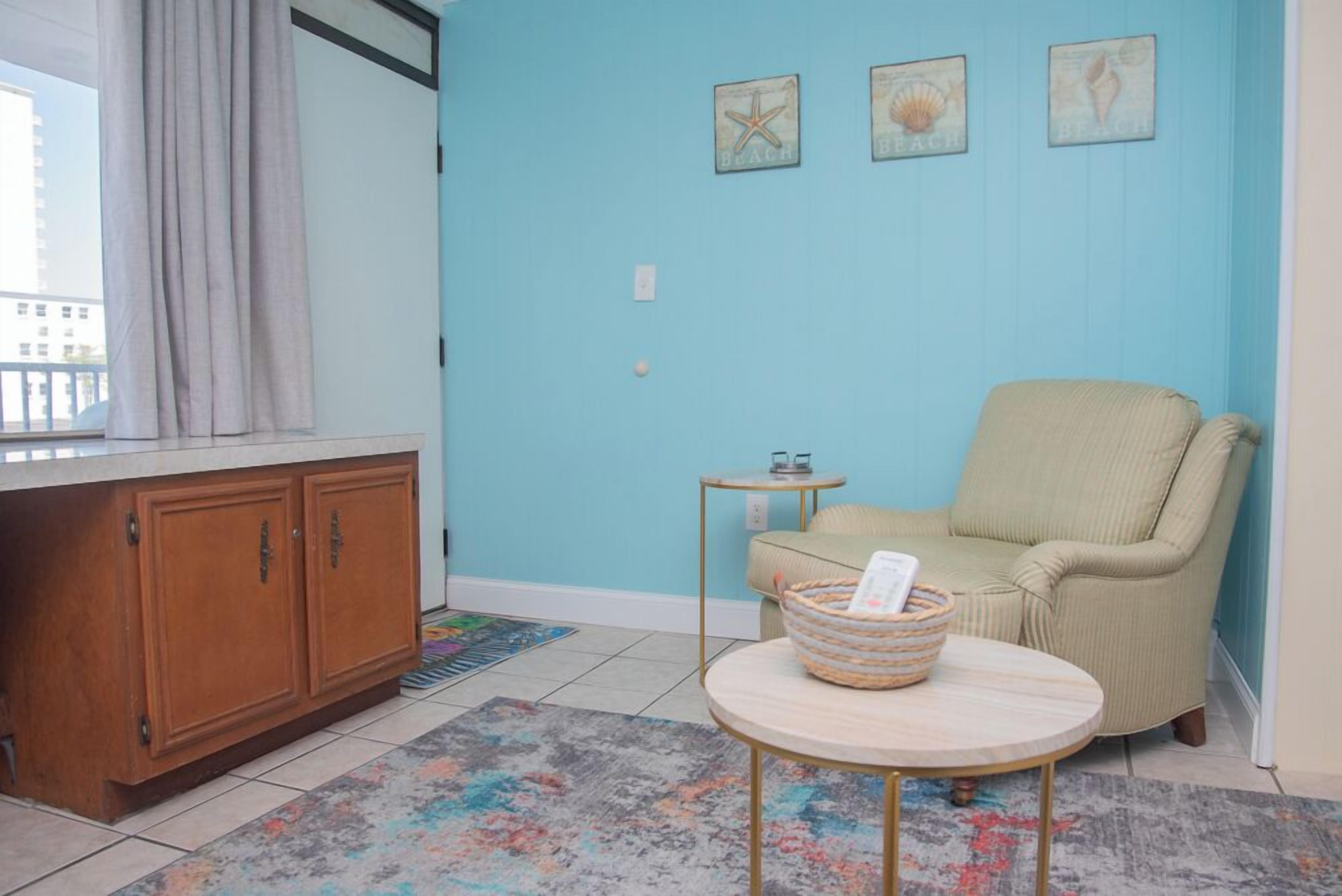 A room with a light blue wall, a wooden cabinet, a beige armchair, a small round table with a basket, and a colorful rug.