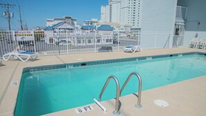 Una piscina con indicador de profundidad de 2.4 metros, rodeada por una valla blanca, con sillas de playa y un puesto de salvavidas al fondo.