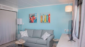 Una sala de estar con pared azul claro, un sofá gris con cojines estampados, una mesita redonda, un tapete colorido y dos cuadros colgados en la pared que representan tablas de surf.