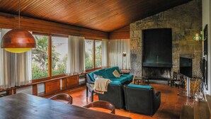 A spacious living area with a stone fireplace, wooden ceiling, and large windows offering a view of greenery.