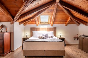 A bedroom with a sloped wooden ceiling, a skylight, a bed with towels, bedside lamps, a wooden wardrobe, and a small table with a lamp. - Iris Stone Built Villa (Agios Nikolaos)