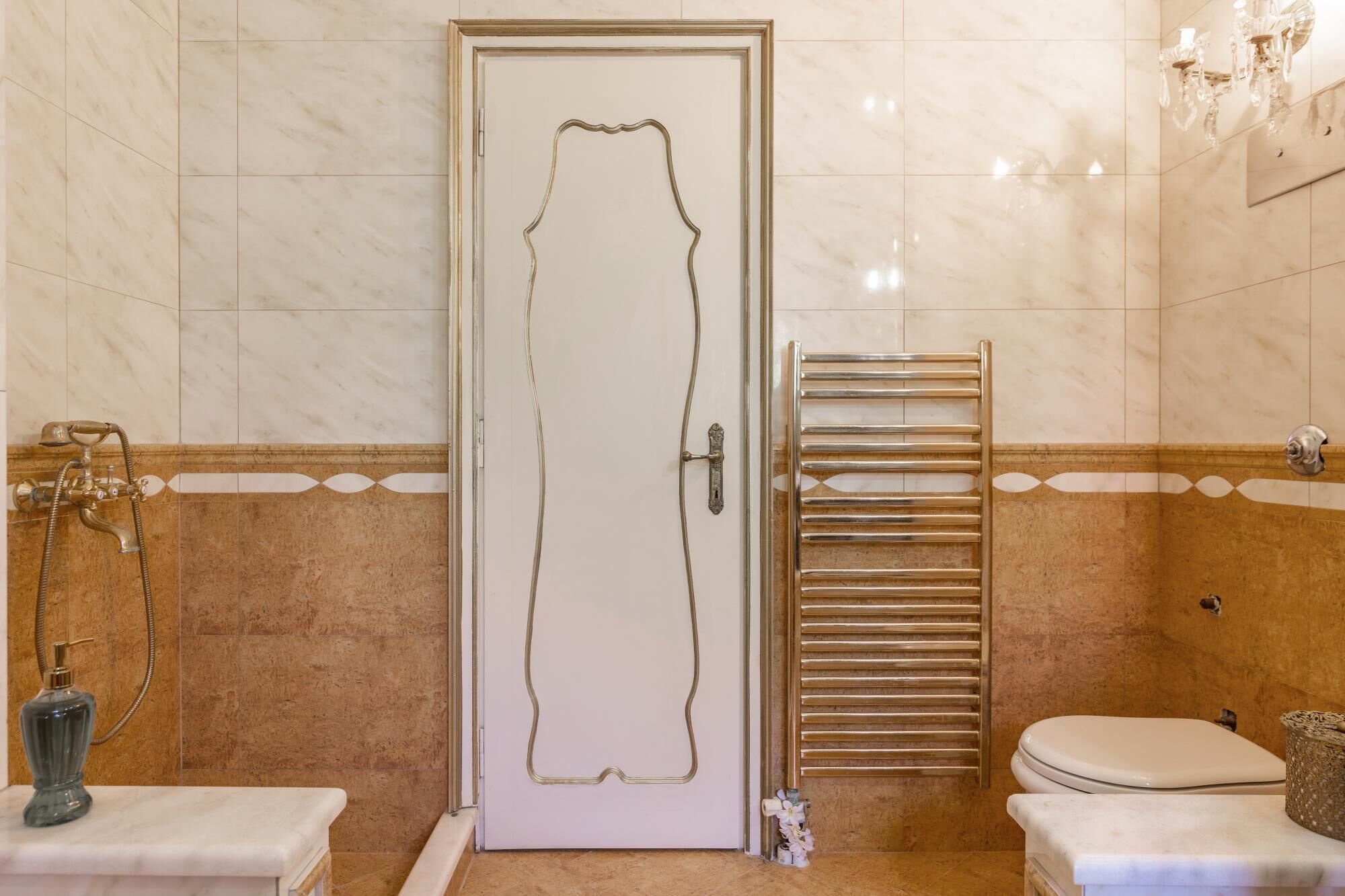 A bathroom with a marble floor, a white door with a gold outline, a shower with a gold faucet, a towel rack, and a toilet.