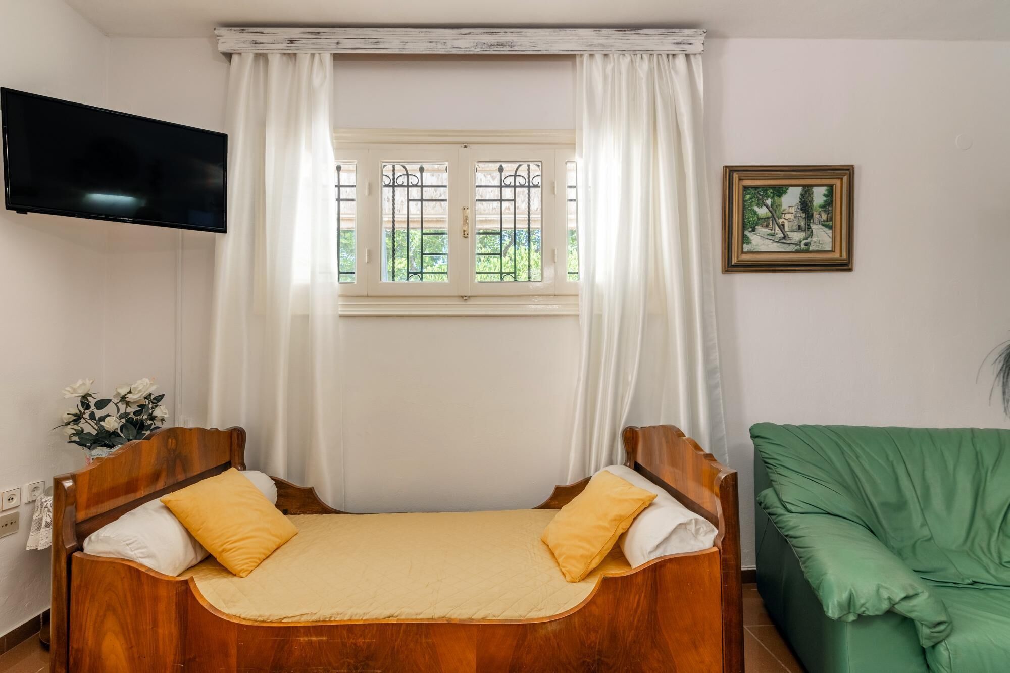 A room with a bed, a green sofa, a TV mounted on the wall, and a window with curtains.