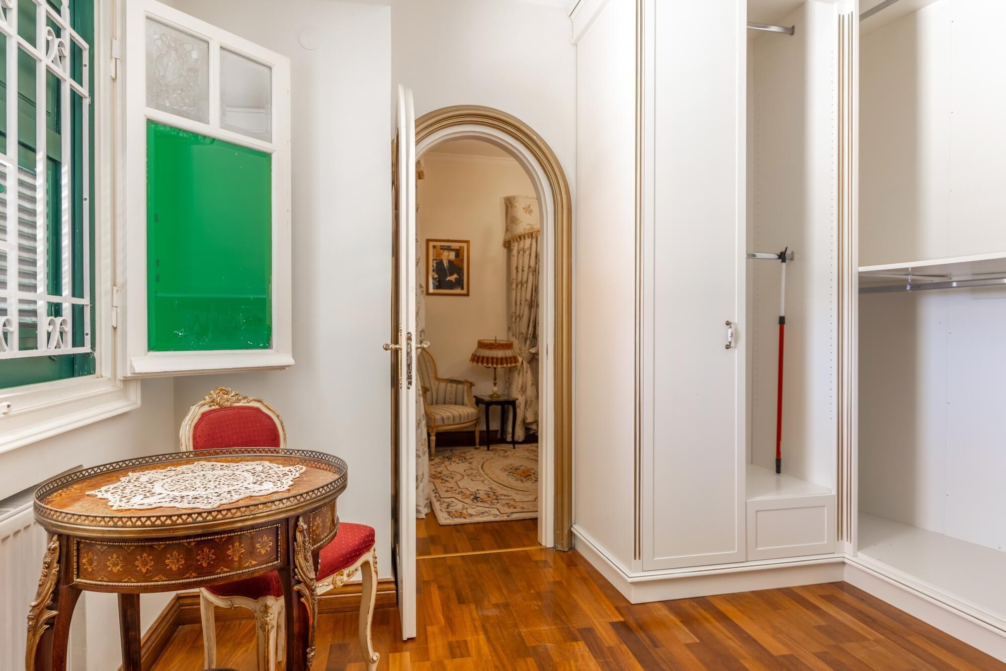 A room with a wooden floor, a round table with a lace tablecloth, a red chair, a green window, and a white wardrobe.
