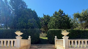 Un escalier en pierre mène à une entrée fermée par un portail, d’où l’on peut admirer un jardin bien entretenu et un ciel d’un bleu éclatant.