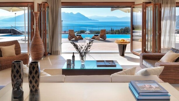 A spacious living room with a glass coffee table, a sofa, and a view of the sea and mountains.