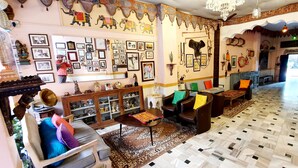 A room with a patterned floor, a wooden cabinet, a table with a decorative cloth, and a wall adorned with framed pictures and wall hangings. - Durag Niwas (Jodhpur)