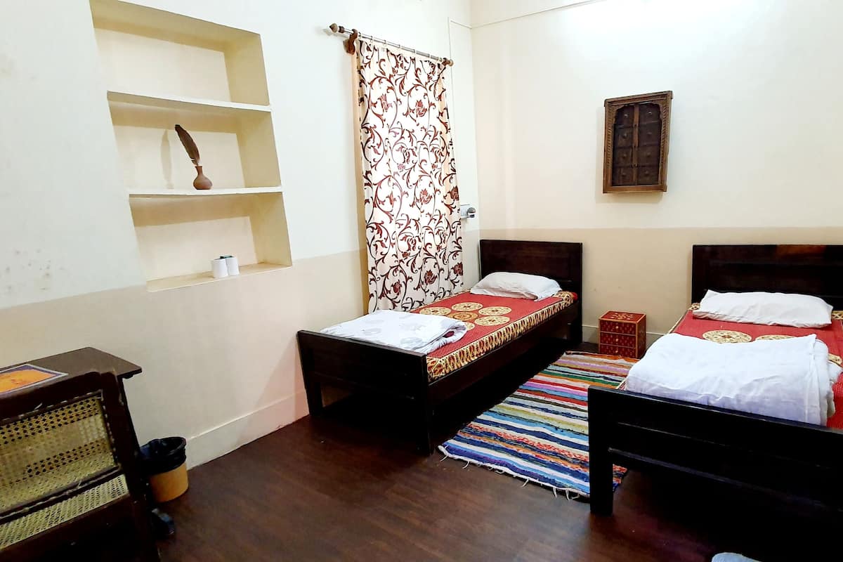 A small, well-lit room with two single beds, a wooden desk, shelves, and a patterned curtain.