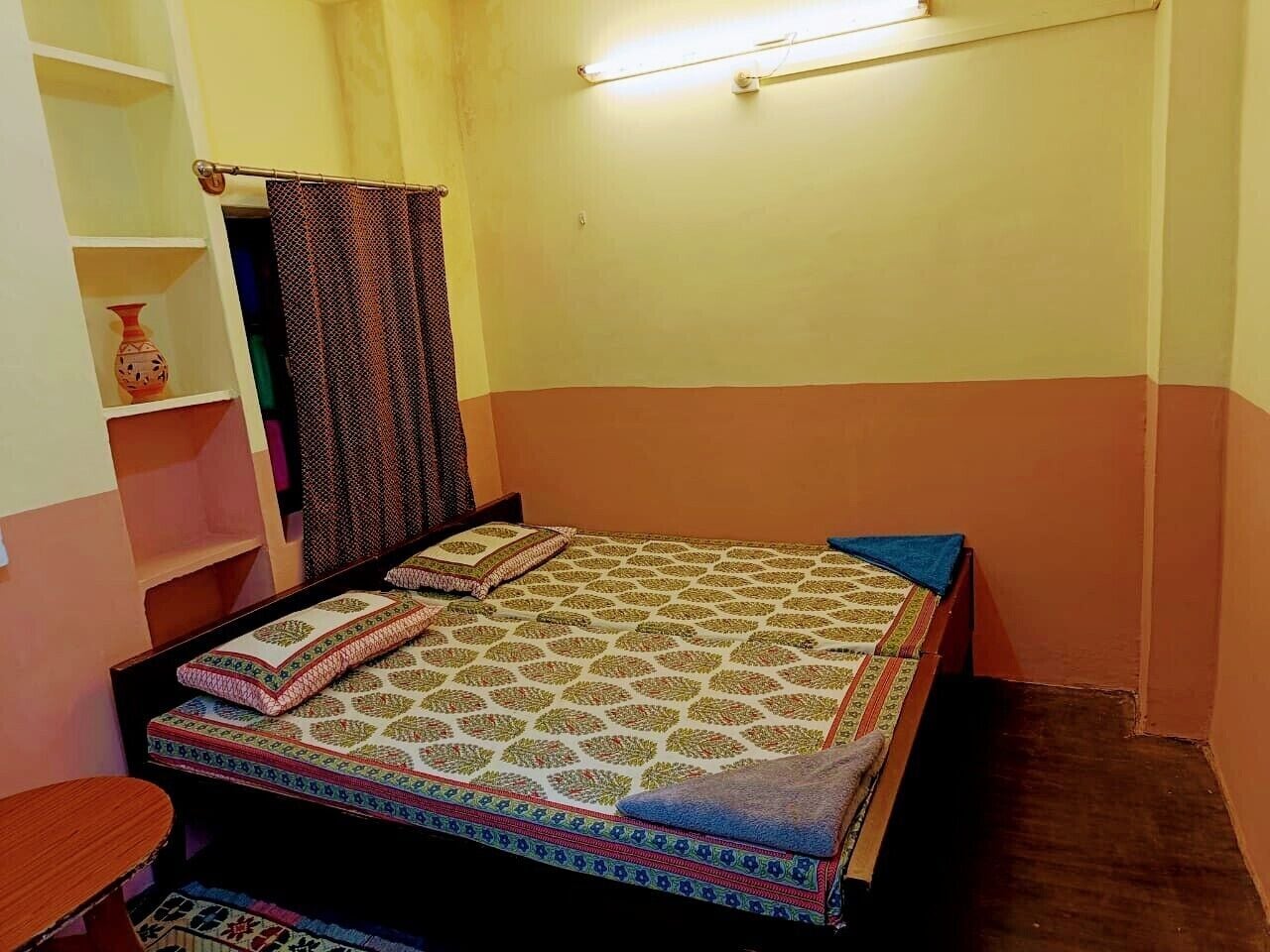 A small, single-bed room with a patterned bedspread, a shelf with decorative items, and a curtain.