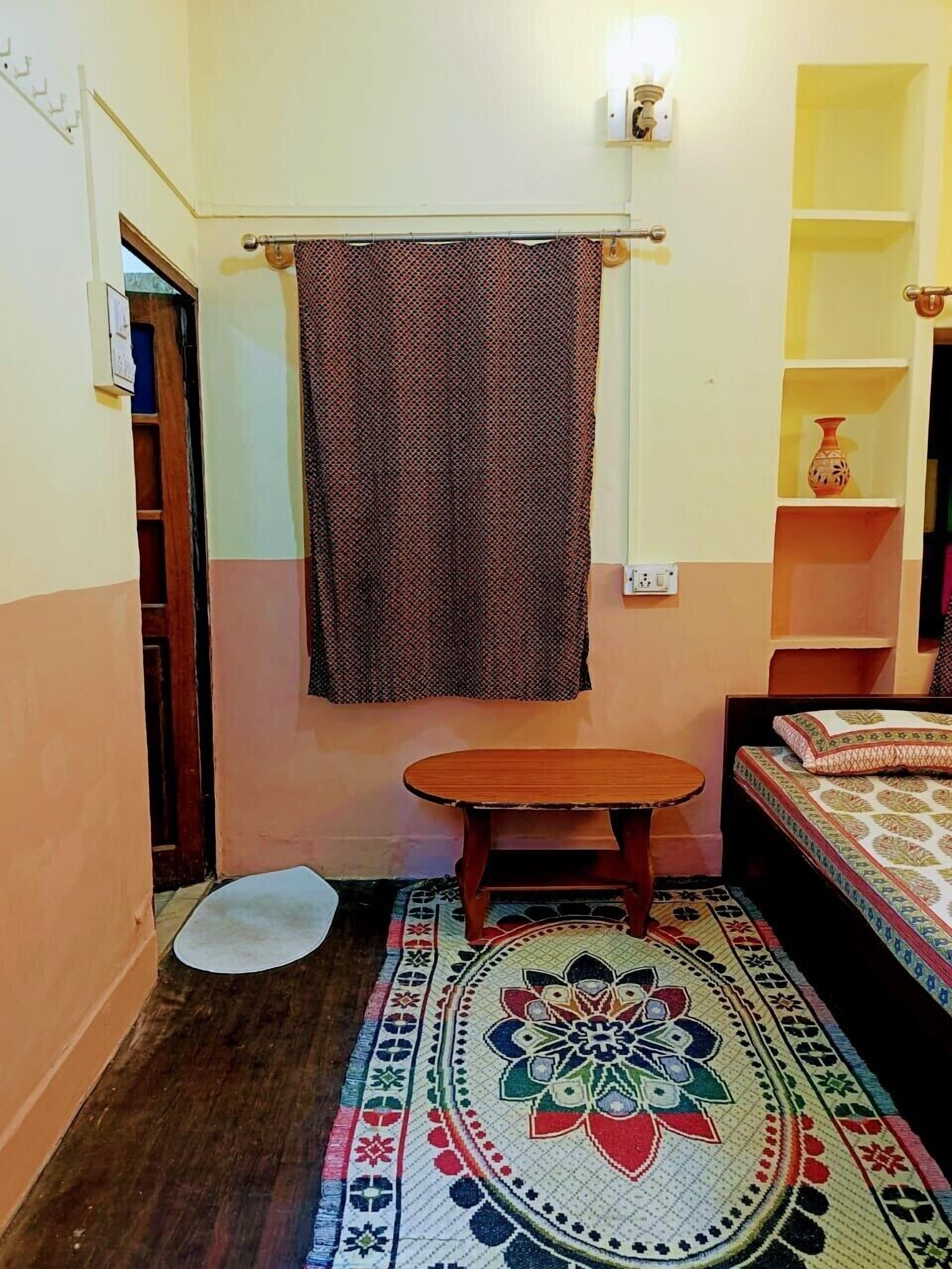 A small, compact room with a single bed, a small table, a patterned rug, and a towel hanging on a rod.
