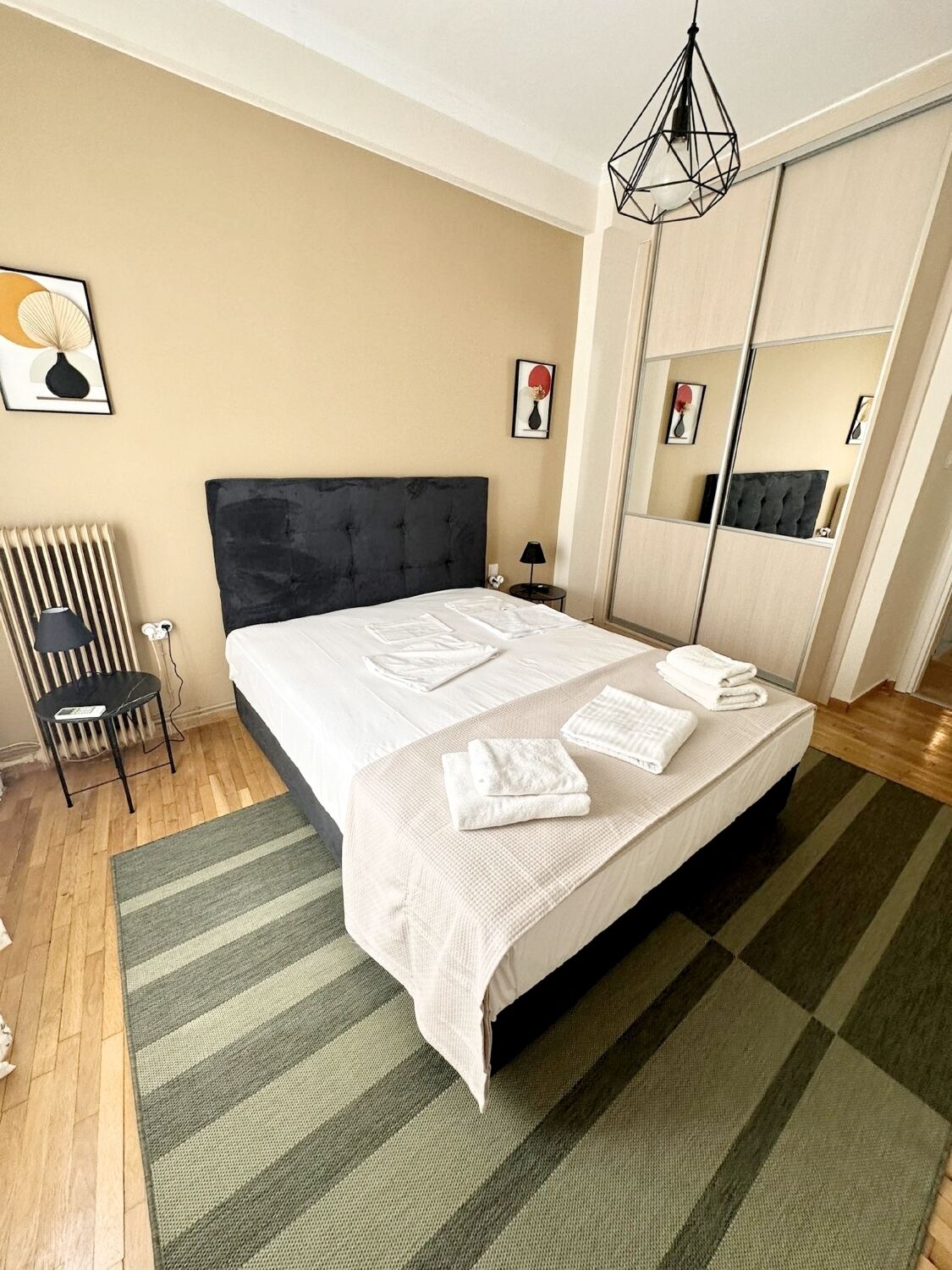 A neatly made bed with white linens and folded towels, a black upholstered headboard, a radiator, a small table with a lamp, and a mirror with a black frame.