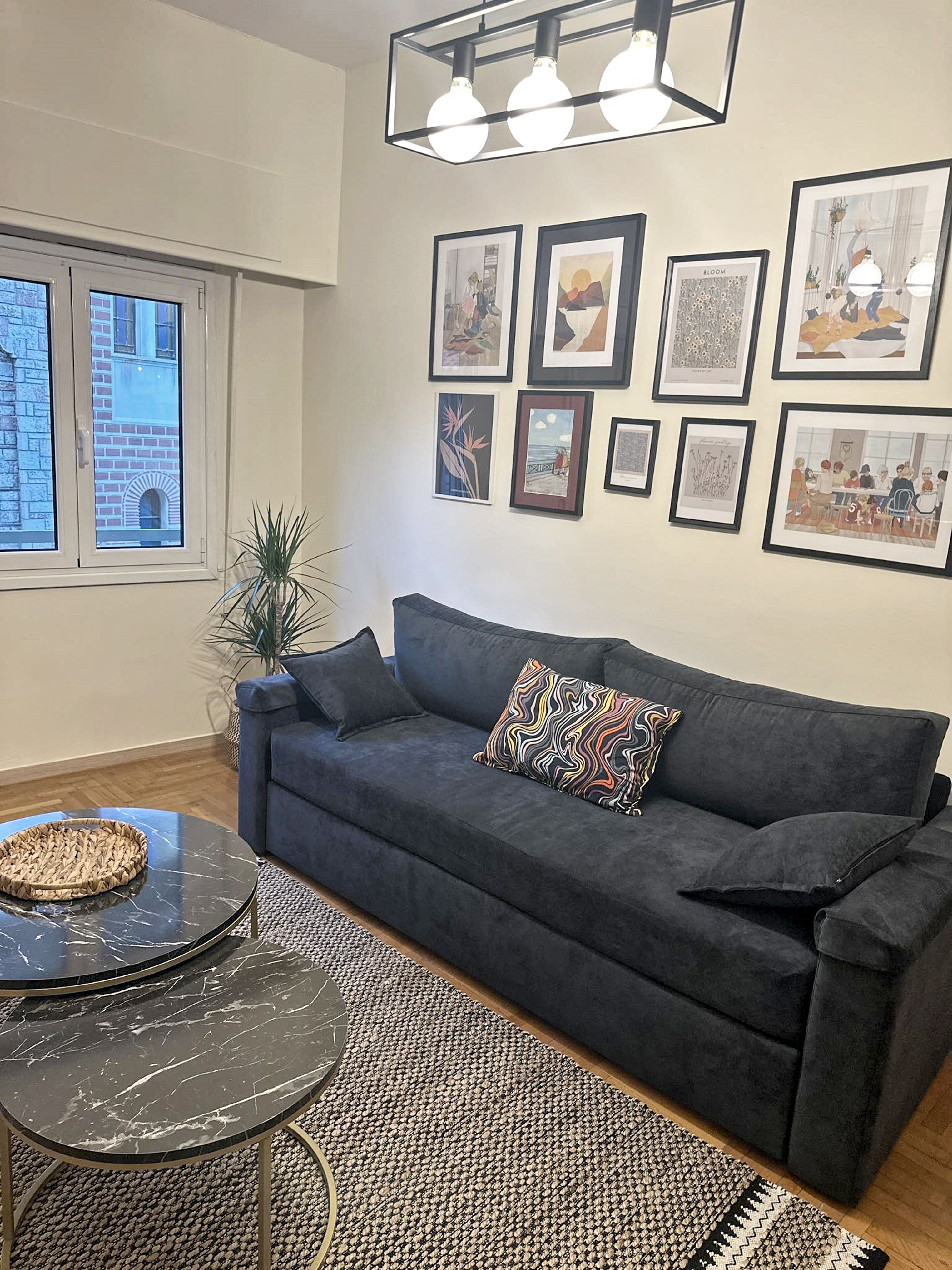 A living room with a dark grey sofa, a round black coffee table, and a patterned rug. There are framed artworks on the wall and a window with a view of buildings.