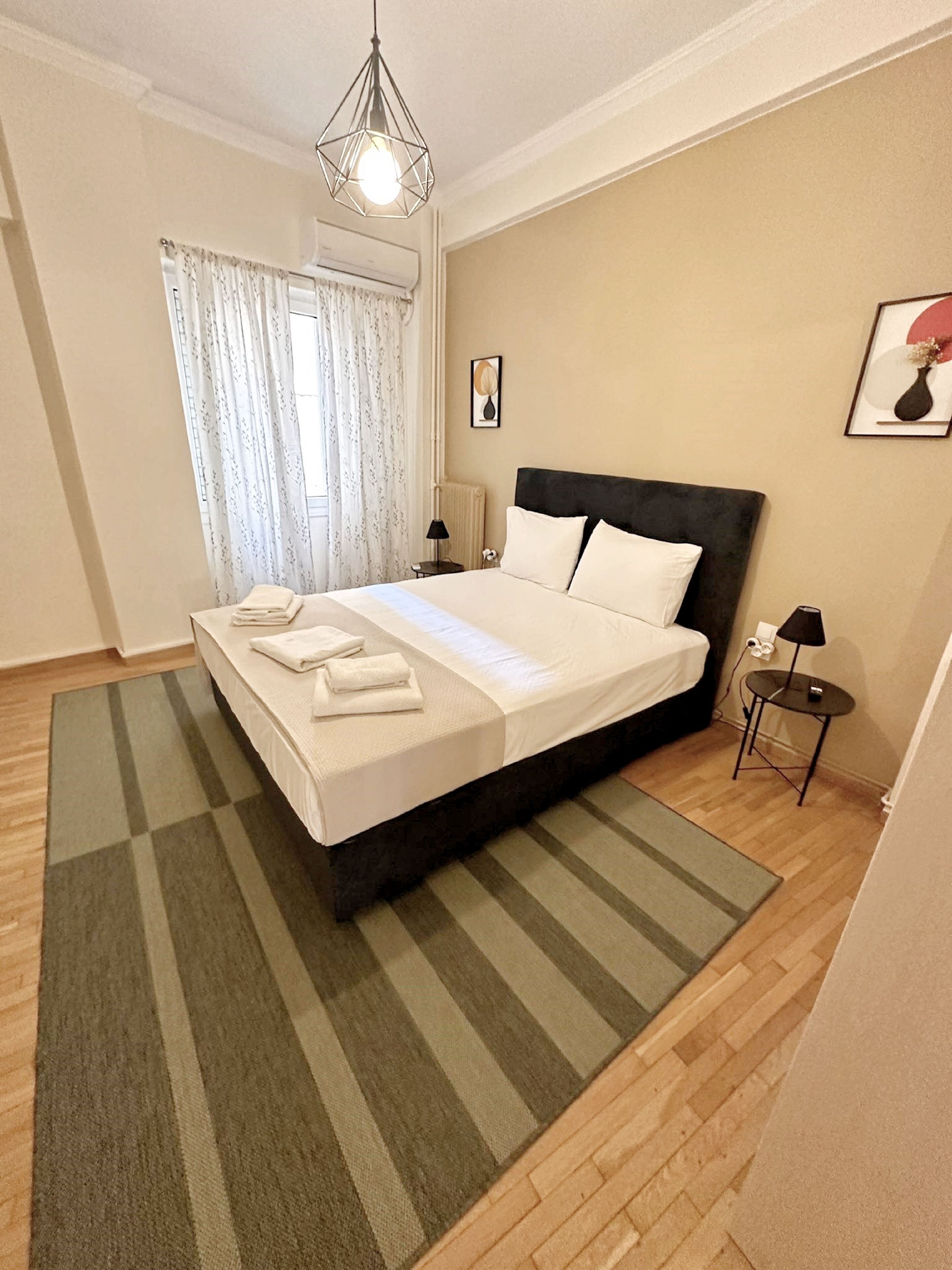 Una cama bien hecha con sábanas blancas y una toalla doblada, una mesita de noche con una lámpara, un cuadro en la pared y una ventana con cortinas.