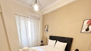 A neatly made bed with white linens and a folded towel, a bedside table with a lamp, a framed picture on the wall, and a window with curtains.