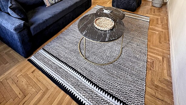 A modern living room with a black sofa, a round coffee table, and a patterned rug. There are framed pictures on the wall and a dining area in the background.
