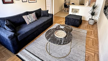 Un salón moderno con un sofá negro, una mesa de centro redonda y una alfombra estampada. Hay cuadros enmarcados en la pared y un comedor al fondo.
