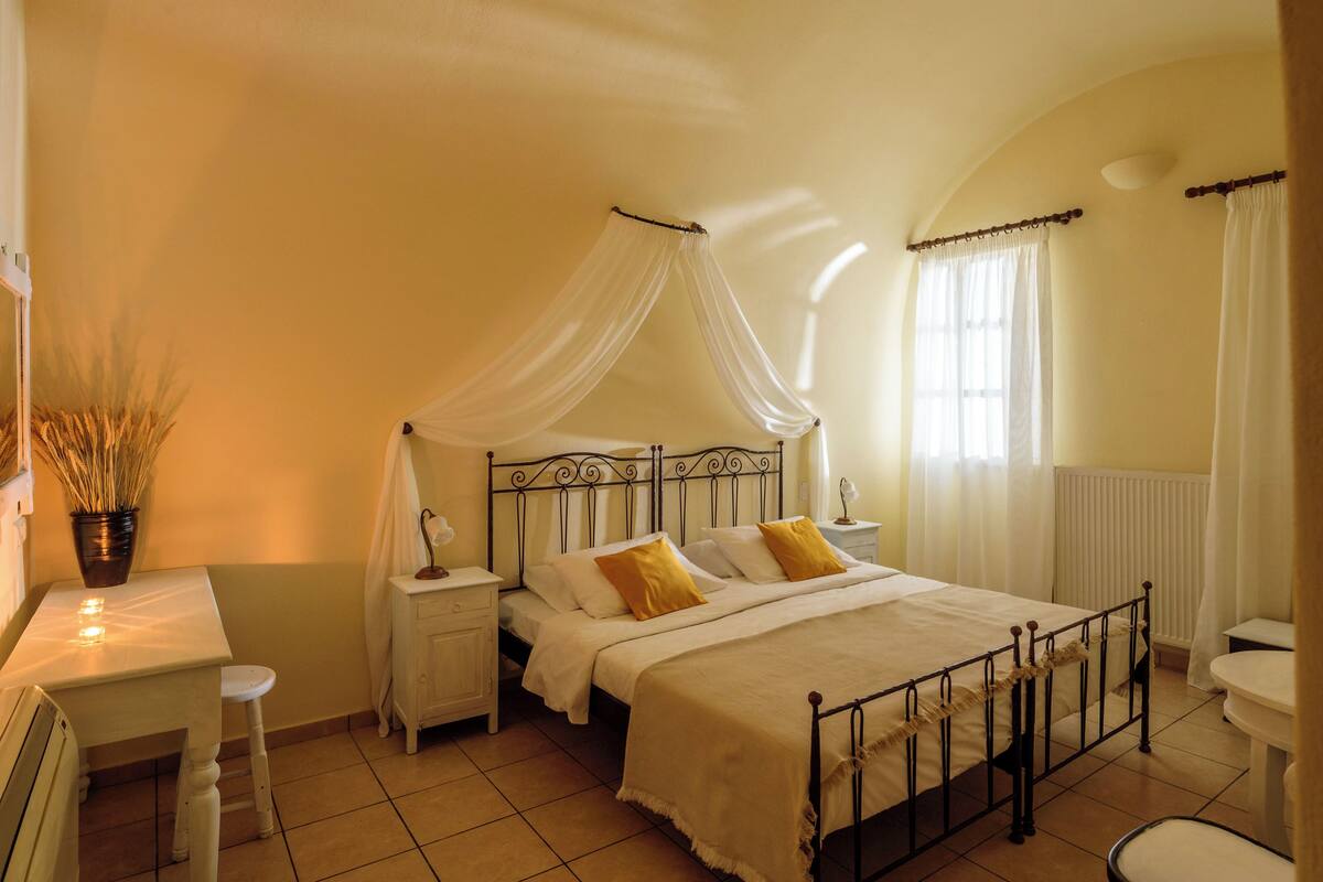 A bedroom with a four-poster bed, a bedside table with a lamp, a chair, and a window with curtains.