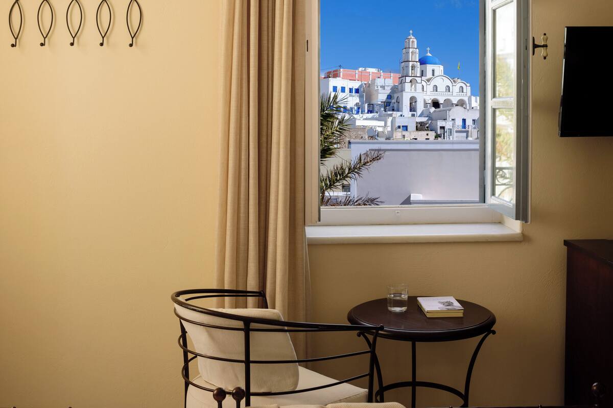 A hotel room with a bed, a small table with a book and glass, a chair, a window with a view of a white building with blue domes, and a curtain.