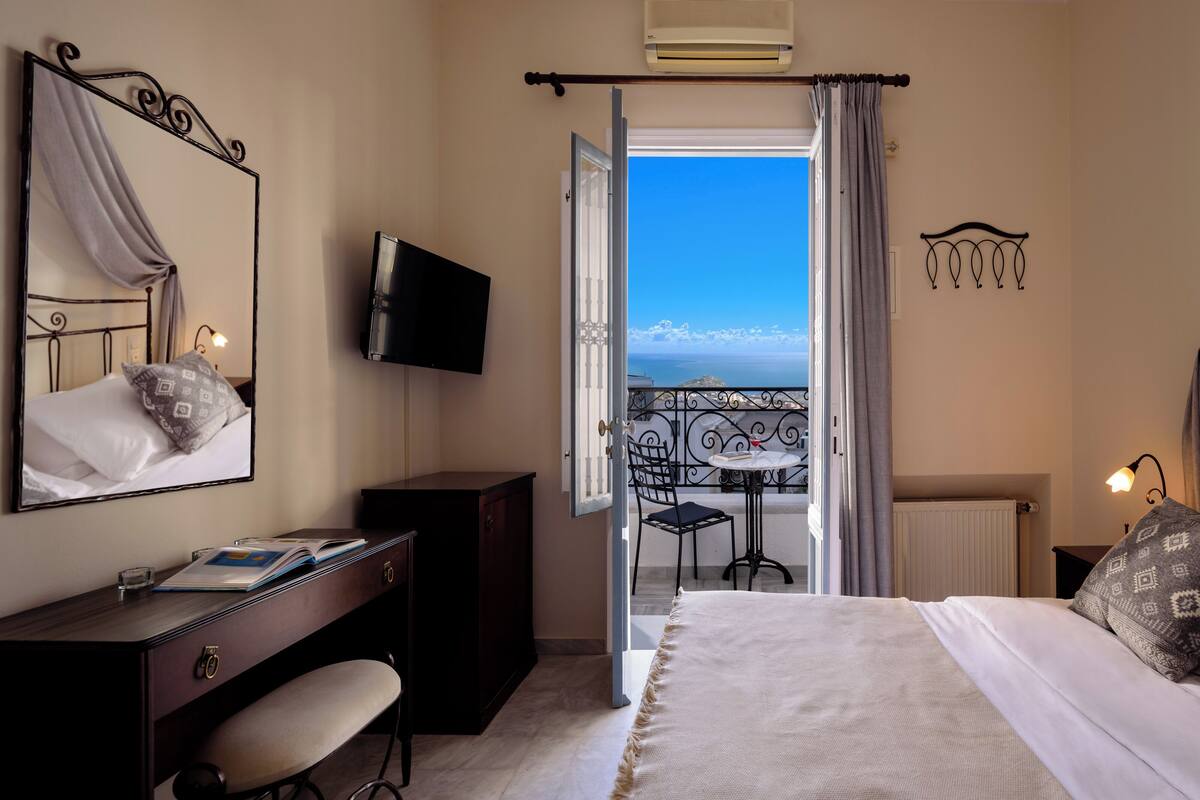 A bedroom with a bed, a desk, a chair, a TV, and a balcony with a view.