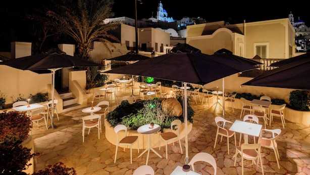An outdoor patio area with tables and chairs under umbrellas, surrounded by stone flooring and plants. - Ikio Concept Santorini (Santorini)