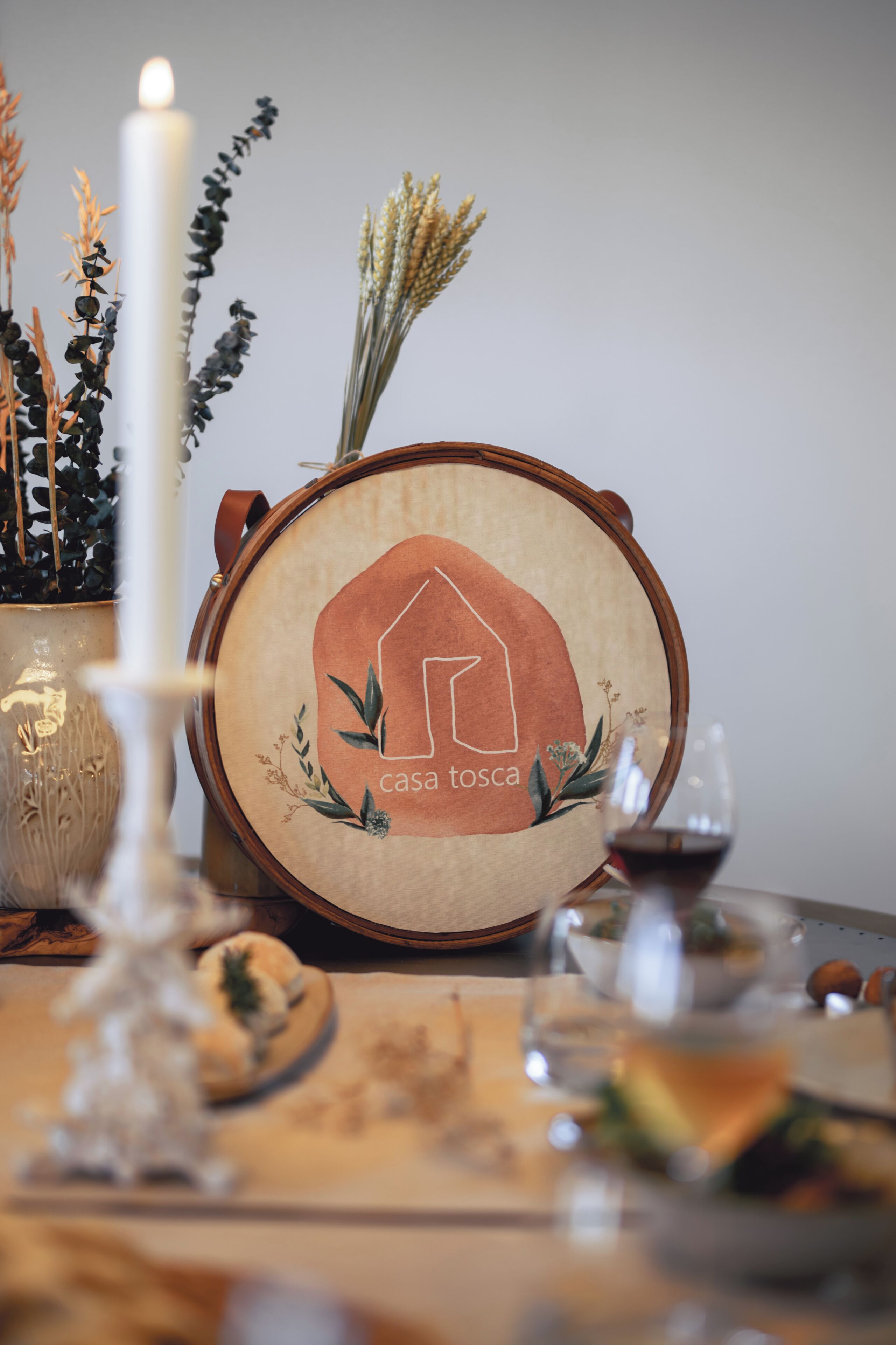 A framed artwork with a house design and the text "casa tosca" on a table with a candle, a glass of wine, and some food items.
