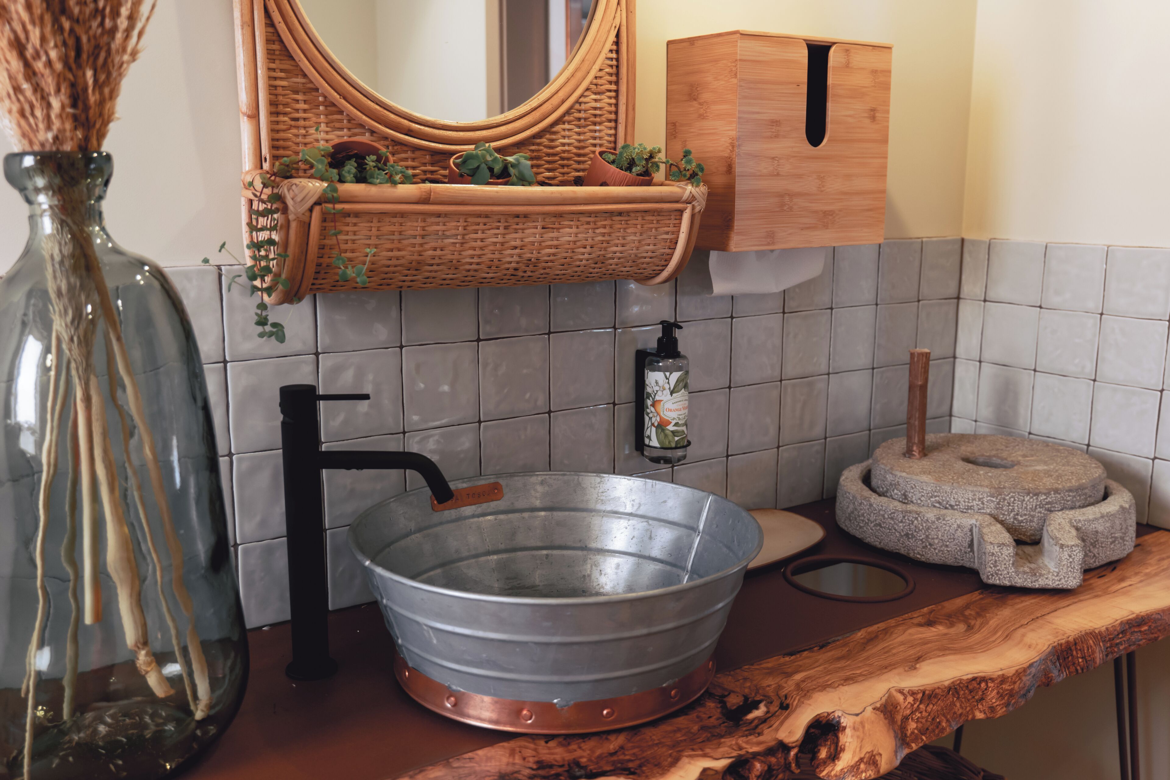A kitchen with a large metal basin, a wooden cutting board, a wicker basket with plants, a mirror, and a wooden cabinet.