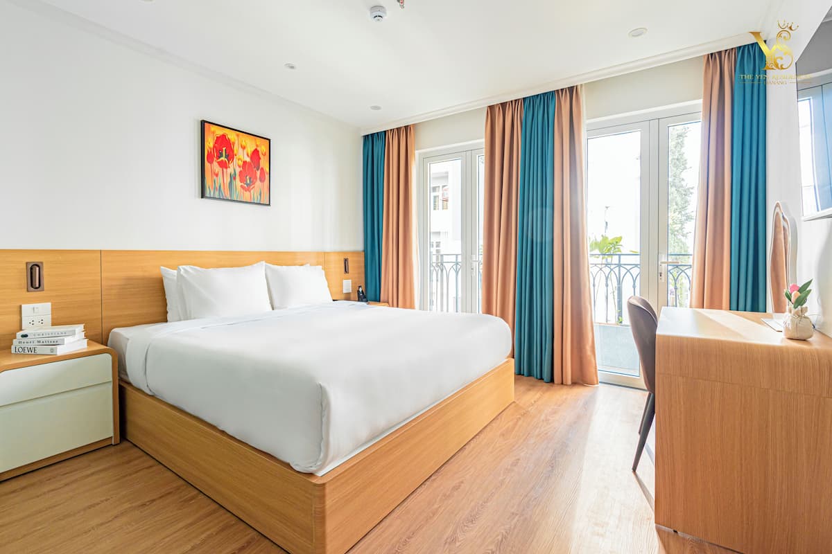 Hotellihuone, jossa on suuri sänky, työpöytä ja tuoli. Seinällä on taulu ja ikkunassa verhot.