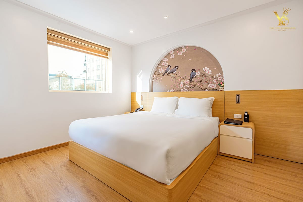 Moderni hotellihuone, jossa on suuri sänky, puinen pääty, yöpöytä ja seinälle kiinnitetty taideteos, jossa on kuvattuna lintuja oksalla.