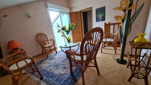 Ett rum med ett matbord, fyra stolar, en blå, mönstrad matta, en gul lampa och ett stort fönster med utsikt över grönområden.