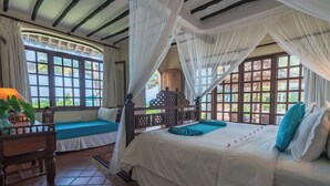 Ein Schlafzimmer mit einem Bett, einem Holzstuhl, einem Tisch mit Pflanze und großen Fenstern mit Vorhängen.