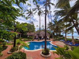 A pool area with palm trees, lounge chairs, and a thatched-roof building.