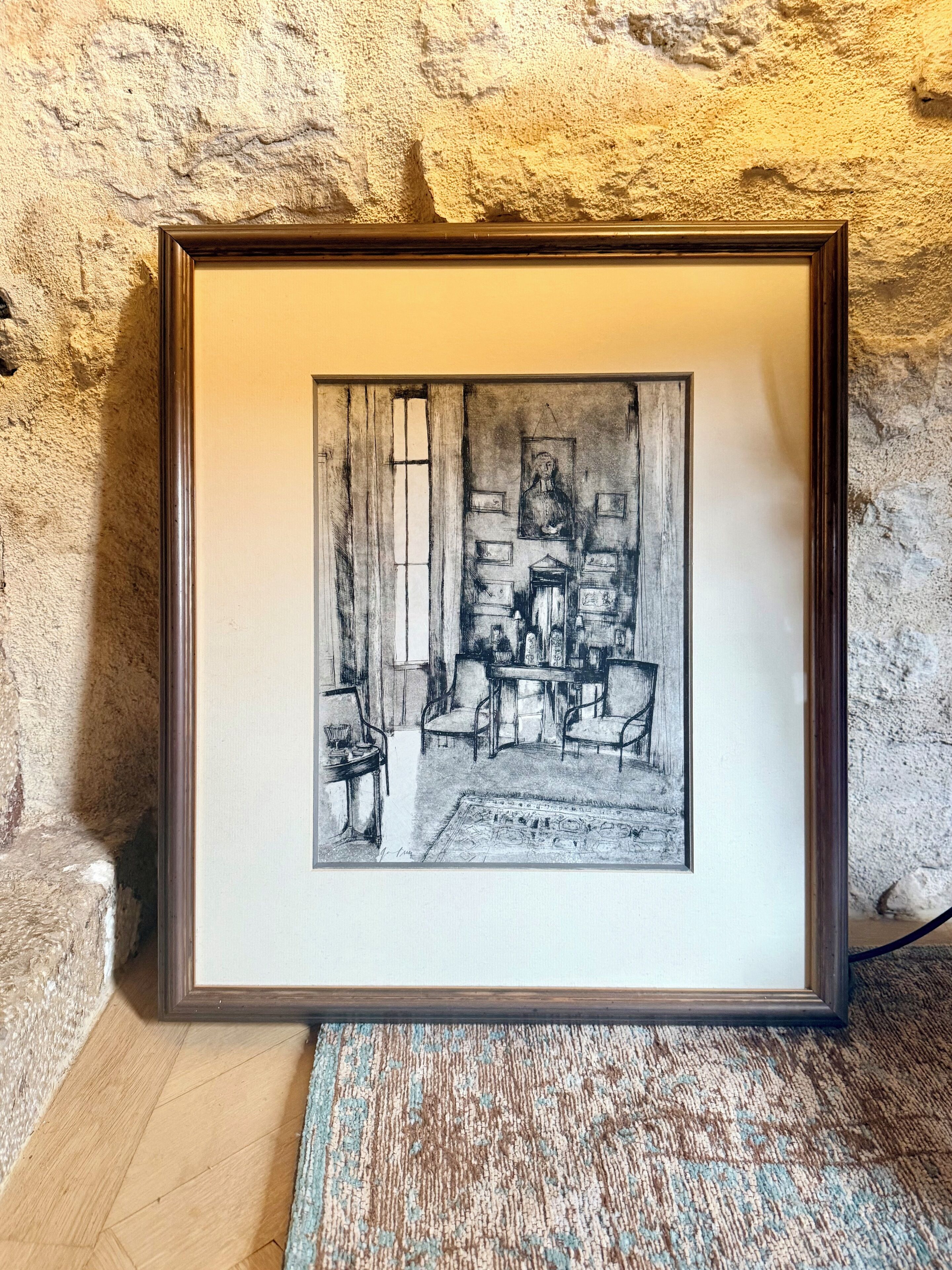 A framed black and white drawing of a room with a table, chairs, and a window.