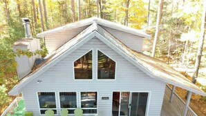 A white cabin with a gabled roof, large windows, and a covered deck with green chairs.