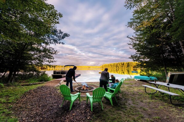 Un camping frente al lago con un hogar, sillas verdes y una mesa de picnic.