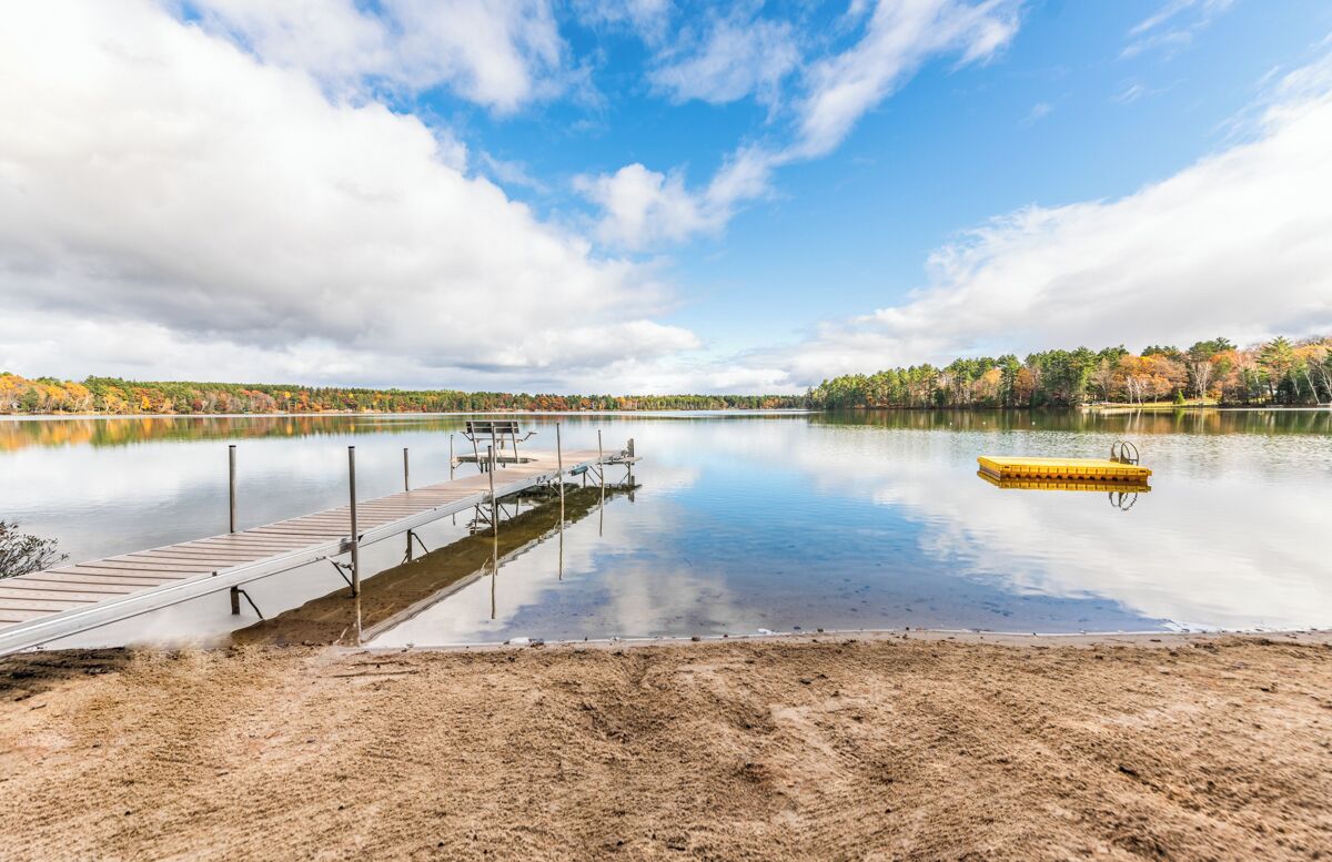 A calm lake with a wooden dock extending into the water, surrounded by trees with autumn foliage.