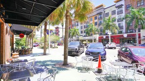 A street with outdoor seating, palm trees, and multi-story buildings.