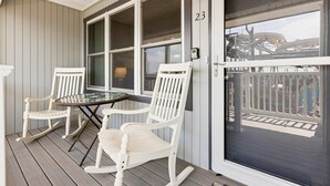 Une véranda avec deux fauteuils à bascule et une table, une porte portant le numéro 23, et une vue sur un pont en arrière-plan.