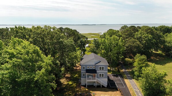 A two-story house with a front porch, surrounded by dense trees and a body of water in the background.