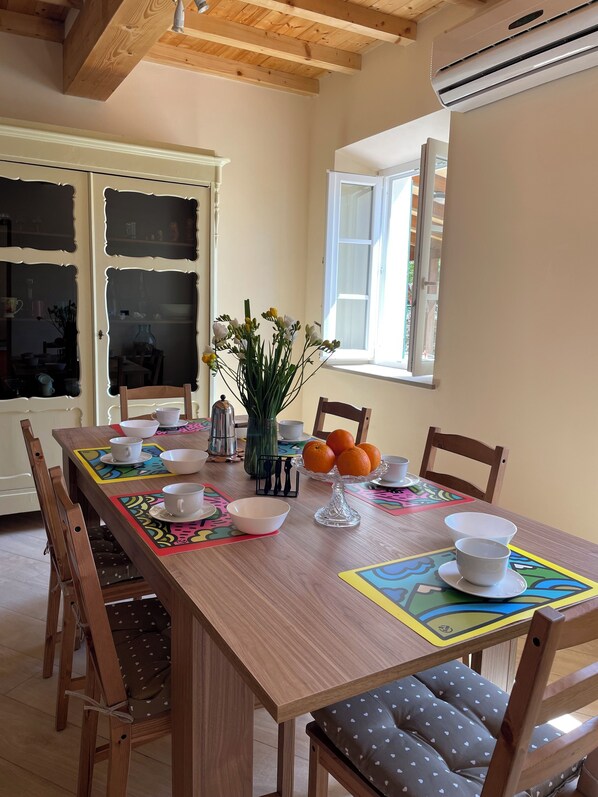 Un comedor con una mesa de madera dispuesta para cuatro personas, rodeada de sillas. Un ventanal proporciona luz natural, y un equipo de aire acondicionado está montado en la pared.