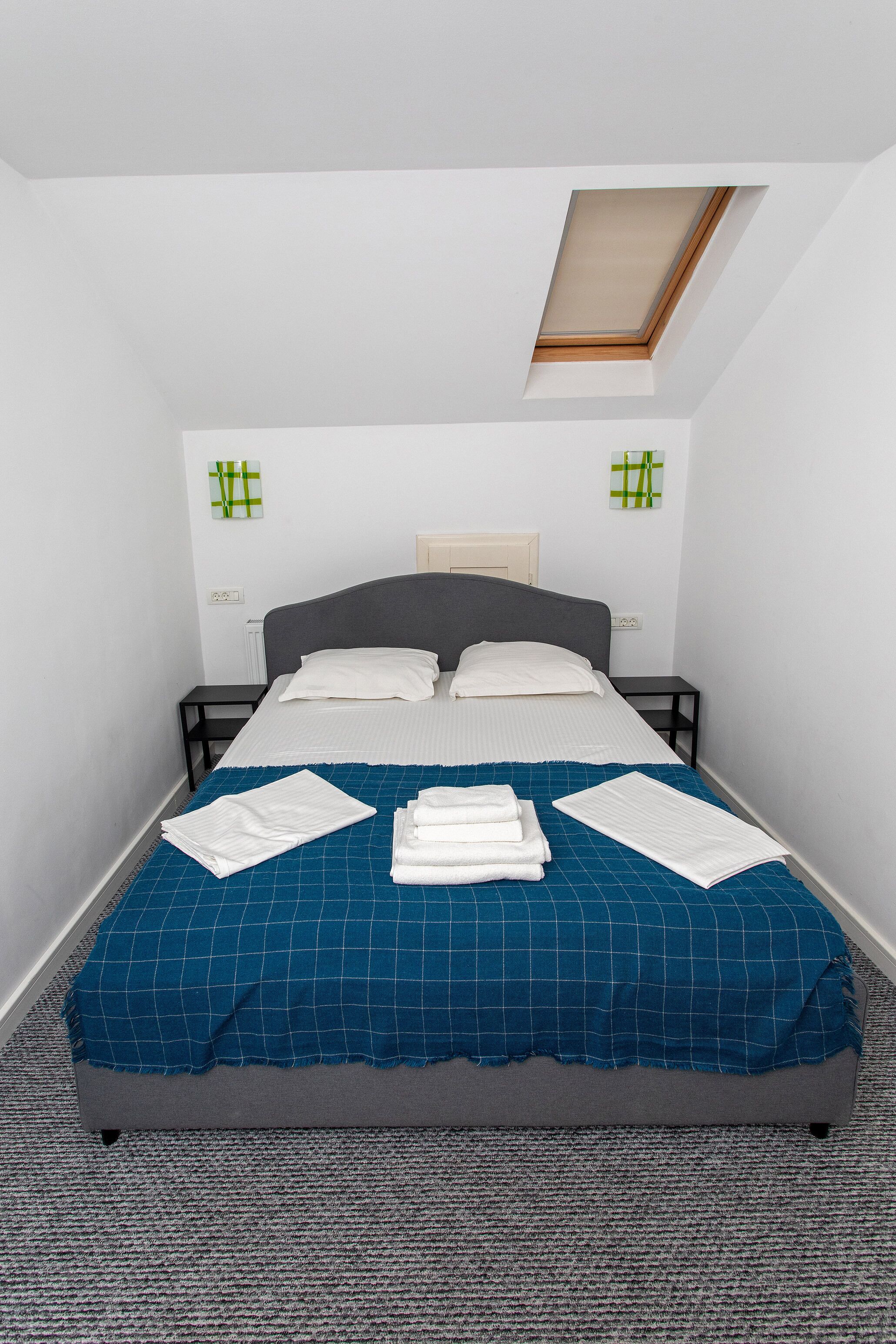 Una cama impecablemente tendida con colcha azul a cuadros, almohadas blancas y toallas blancas dobladas. Hay un tragaluz sobre la cama y la habitación tiene paredes blancas con dos adornos de pared blancos y verdes a cuadros.
