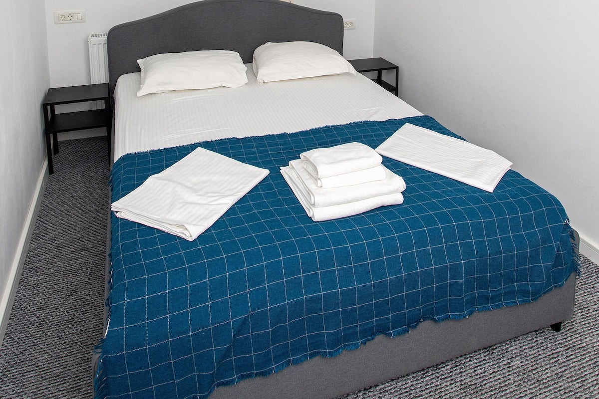 Cama bem arrumada com colcha azul quadriculada e almofadas brancas, uma pequena mesa de cabeceira e um claraboia acima.