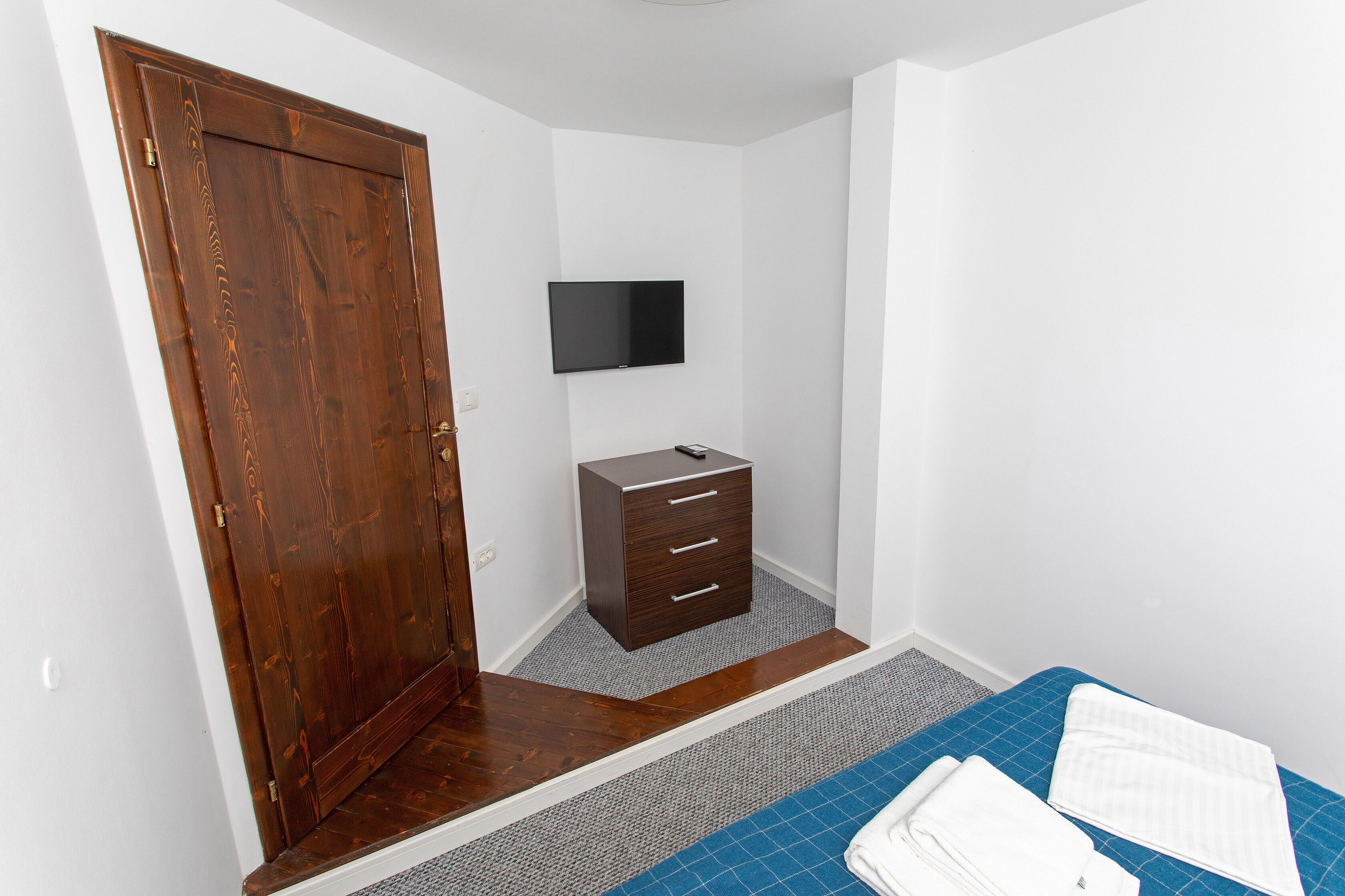 A bedroom with a wooden door, a small dresser, a flat-screen TV mounted on the wall, and a bed with a blue mattress and white bedding.