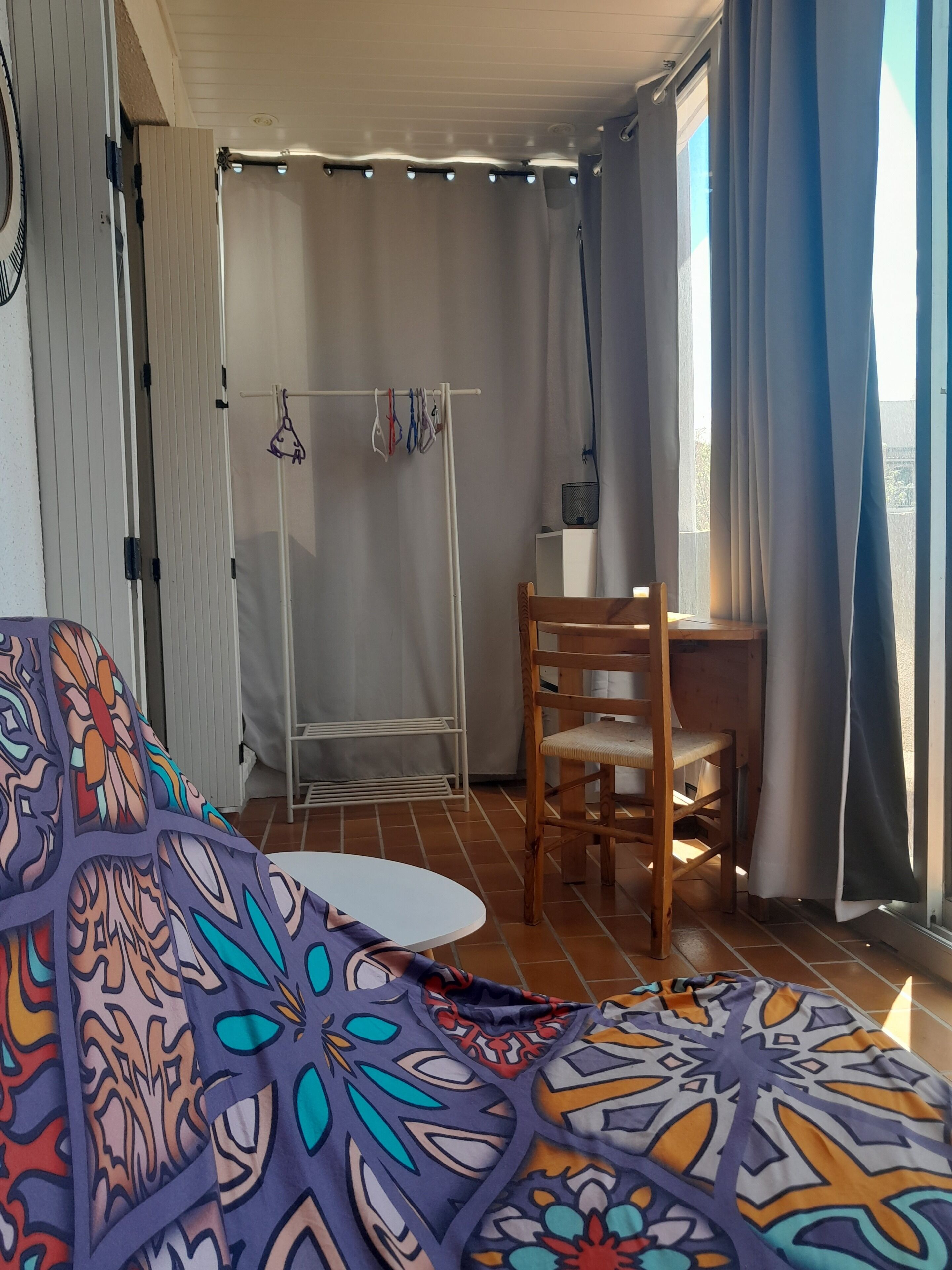 A room with a patterned bedspread, a wooden chair, a small table, and a window with curtains.