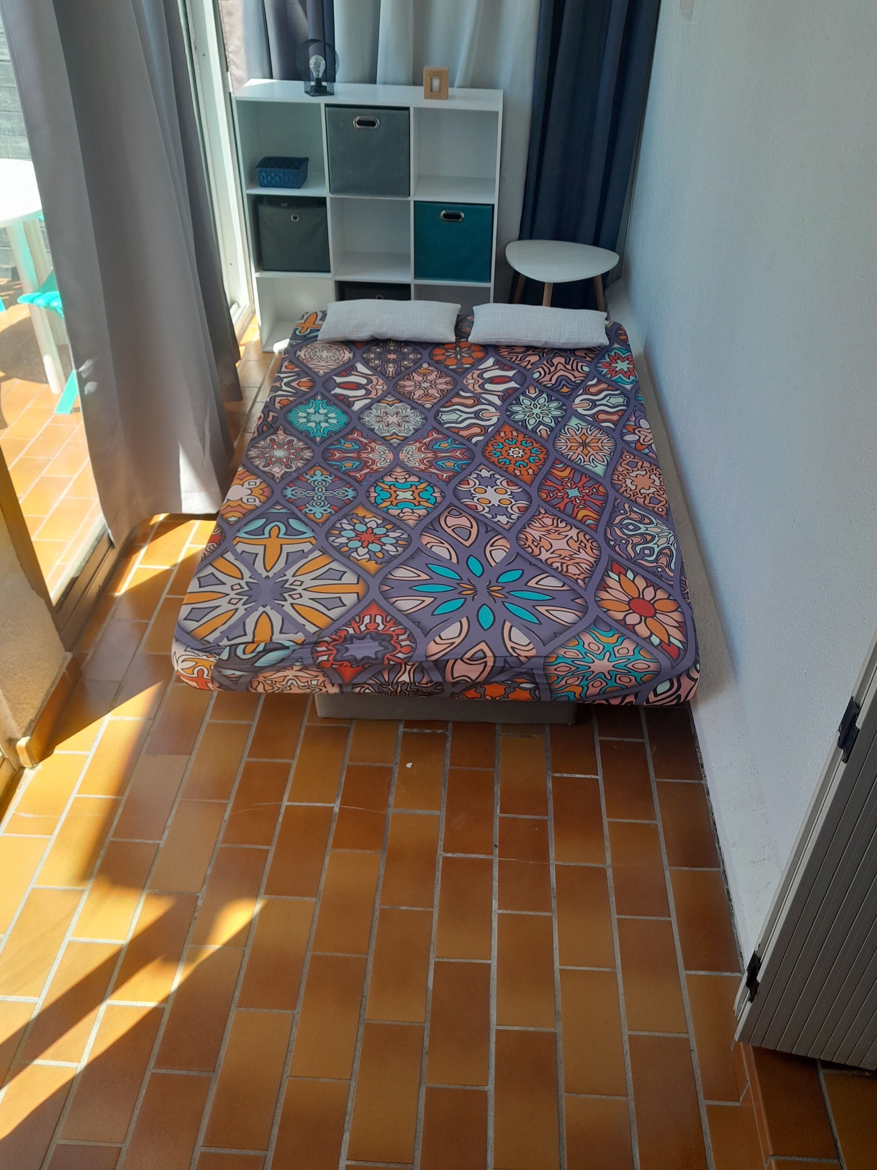 Een kleine kamer met een bedovertrek met patroon, een witte schapenkast en een betegelde vloer.