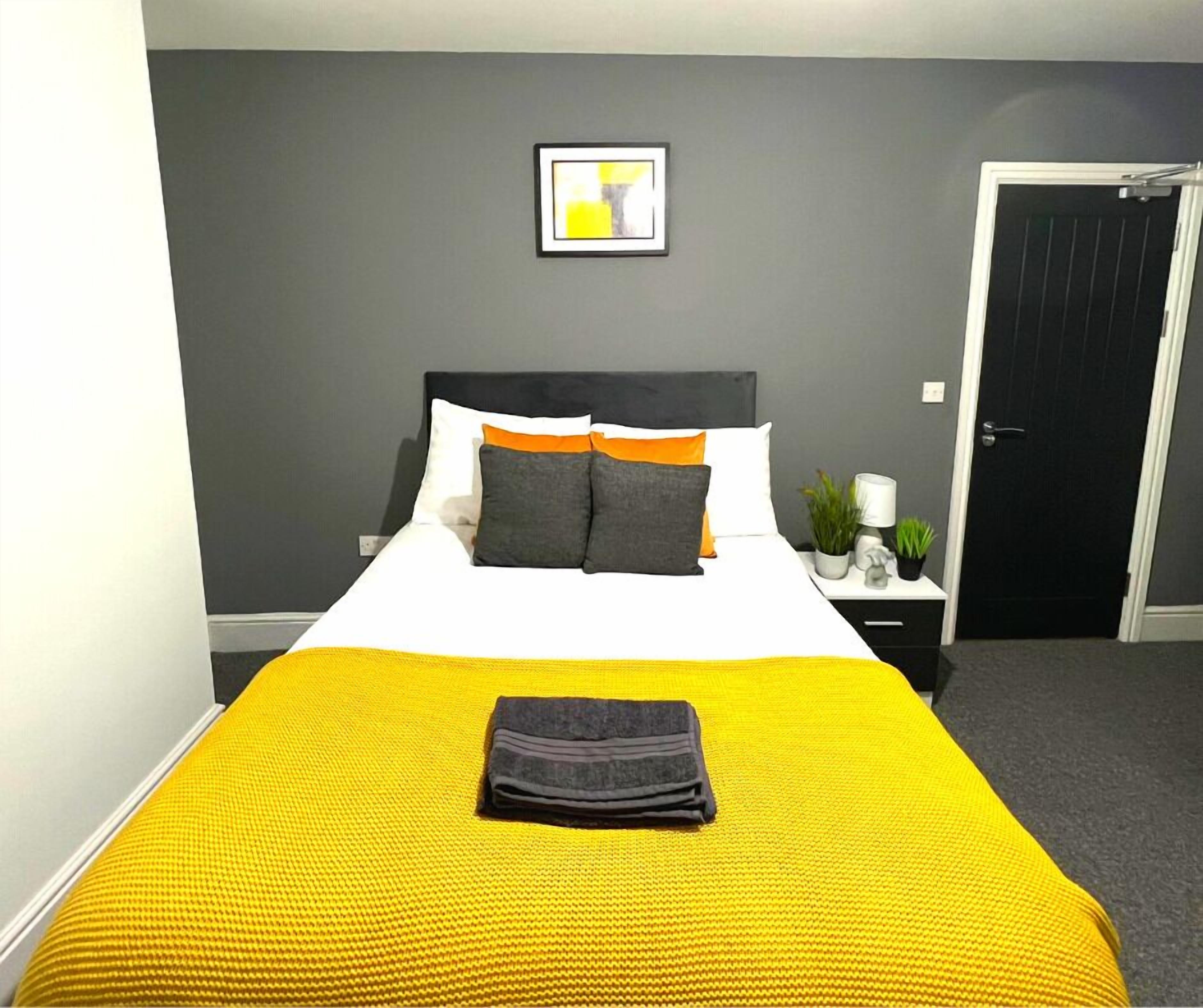 A neatly made bed with a yellow bedspread and a folded towel, a bedside table with a lamp and plants, and a door to the right.