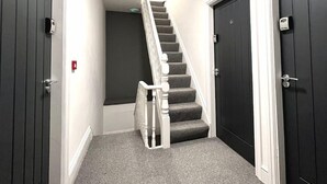 A hallway with a staircase leading to a dark room, a door with a number, and a fire alarm on the wall. - Chantry Suites by SA Stays (Grimsby)